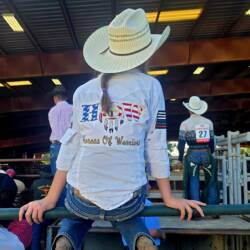 Young lady in white cowboy hat and Horses of Warriors western shirt sitting on metal fencing with back to the camera. She's looking into an arena along with others and cowboys waiting their turn.