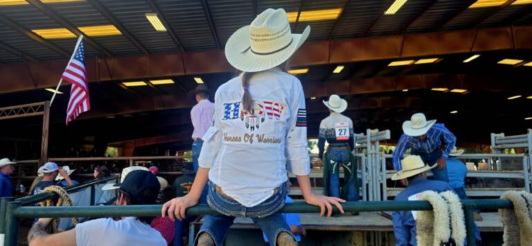 Young lady in white cowboy hat and Horses of Warriors western shirt sitting on metal fencing with back to the camera. She's looking into an arena along with others and cowboys waiting their turn.