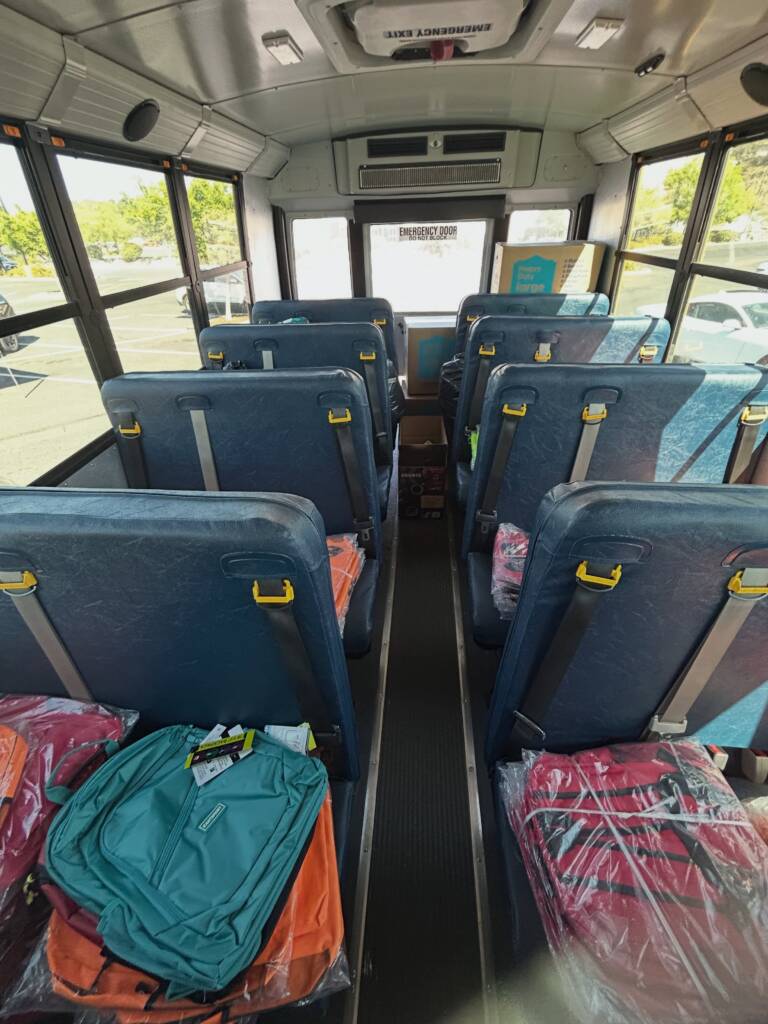 View looking inside a school bus. The seats are filled with new backpacks and school supplies.